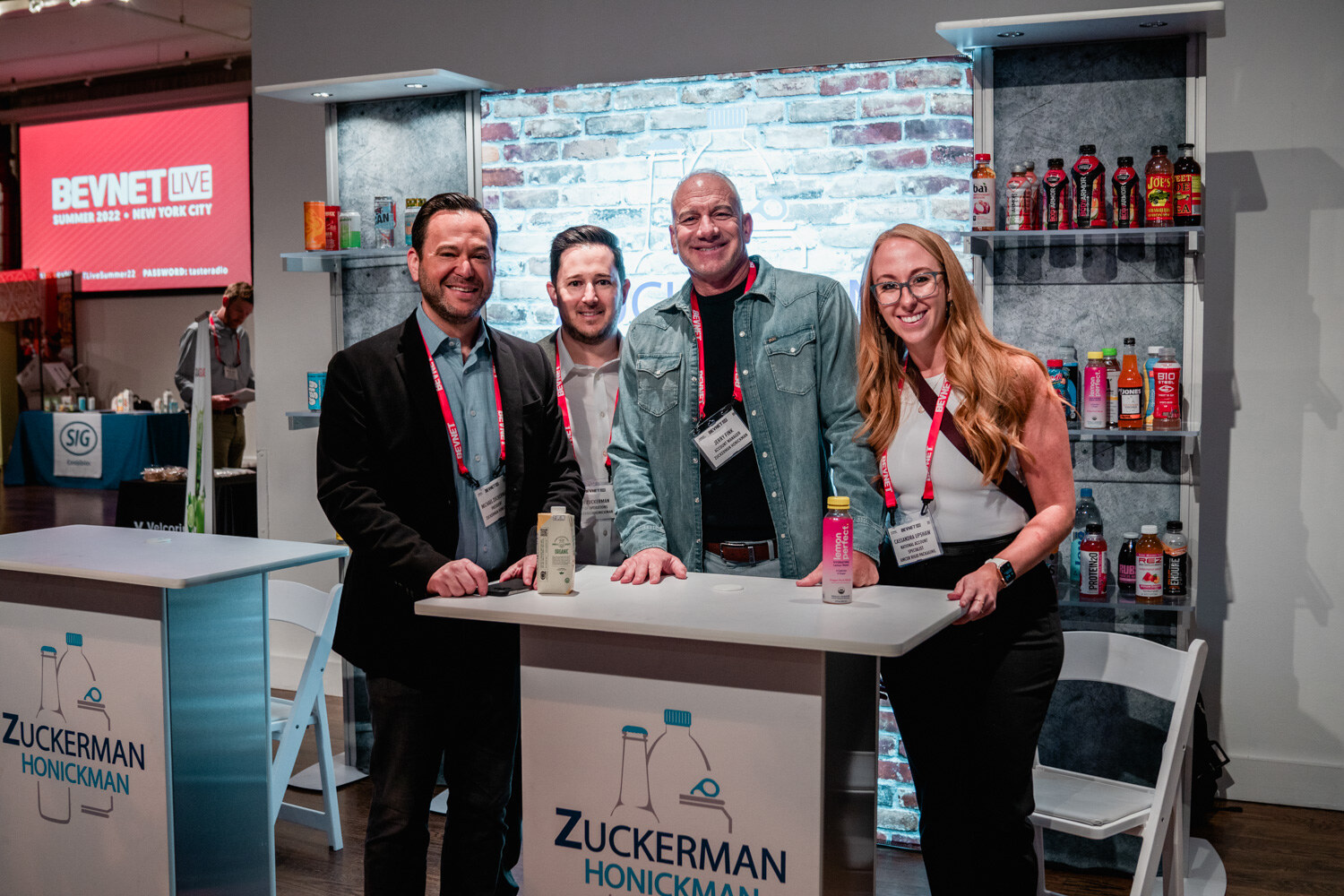 Two men in jackets, a man in a denim shirt and a woman in a white top smile at a standing table in front of a brick wall display with drinks.