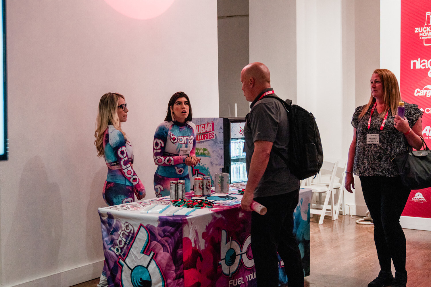 Two women in pink and blue Bang costumes stand behind a table with a Bang tablecloth while two attendees talk to them.