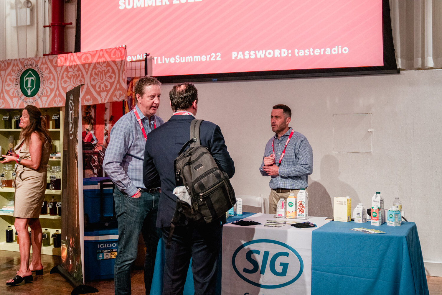 Three men talk beside a blue SIG tablecloth with cartons on it, a screen above reads Password tasteradio.