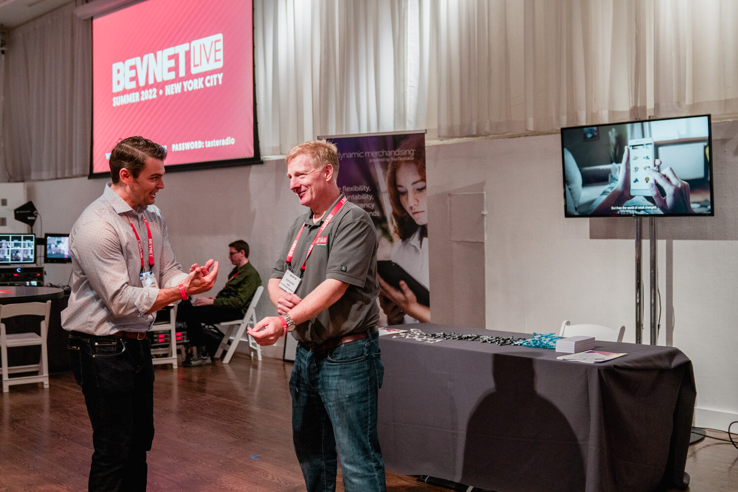 A man in a gray shirt and a man in a gray polo gesture while speaking, a screen and a table behind them.