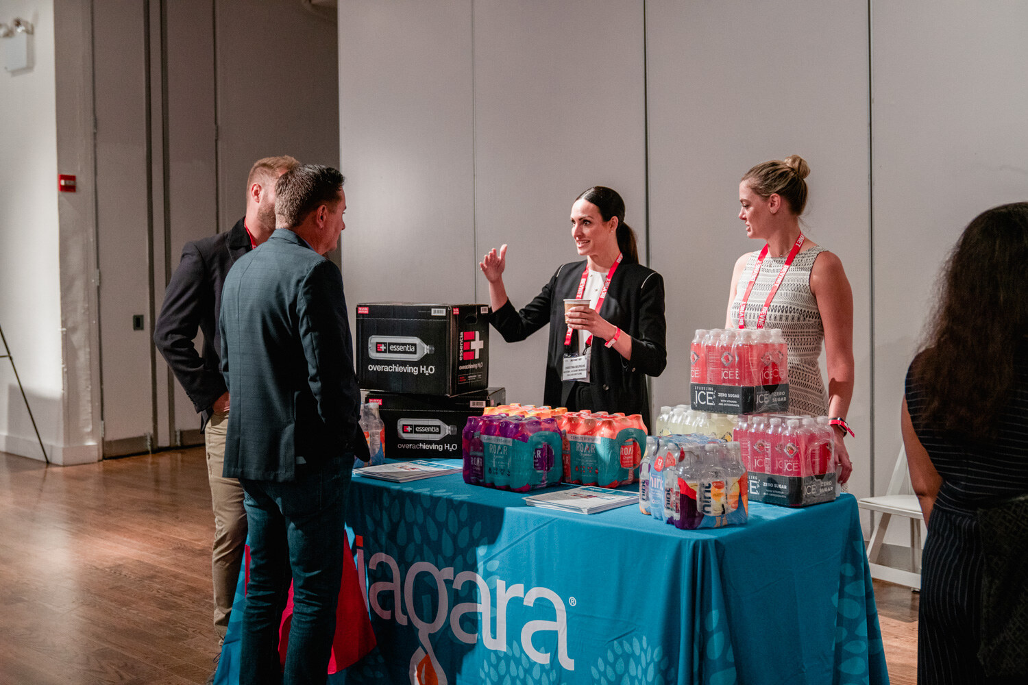 A woman in a black jacket gestures to a group of attendees at a teal Niagara tablecloth stacked with bottled drinks.