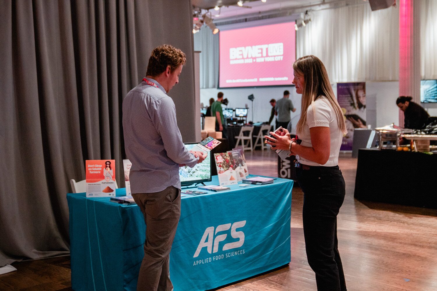 A man in a blue shirt looks at a tablet while a woman in white talks to him at a teal Applied Food Sciences table.