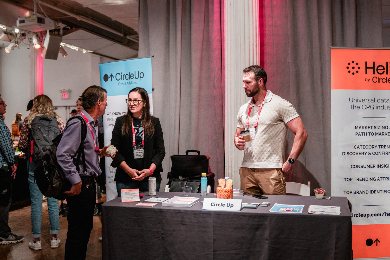 A woman in black and a man in a cream polo stand at a table with CircleUp signage and a Helio by CircleUp banner.