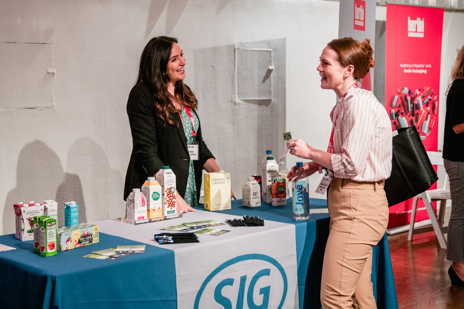 Two women laugh and talk over a SIG tablecloth with juice and milk cartons and a red Berlin banner behind.