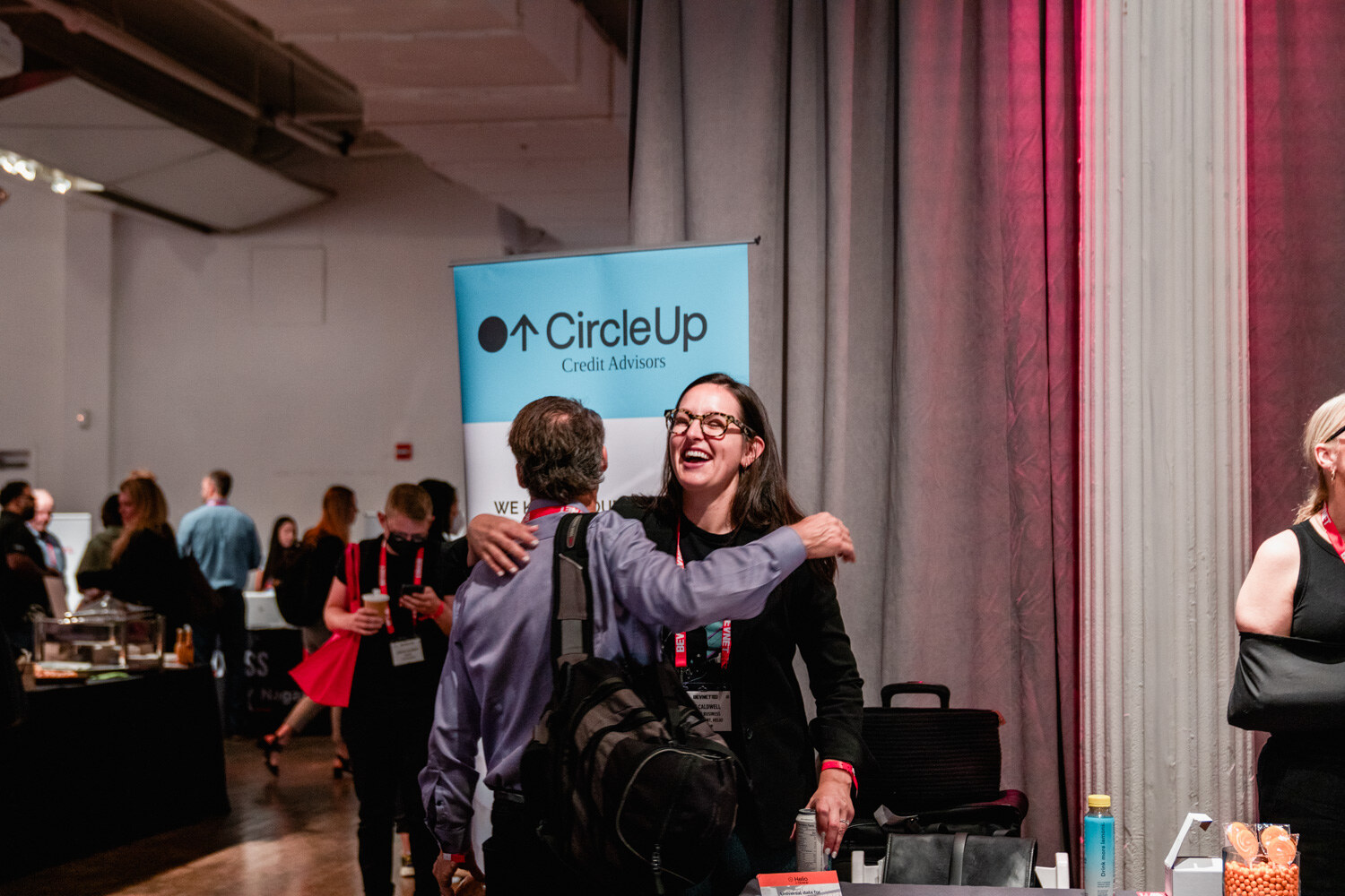 A man with a black backpack hugs a smiling woman in glasses in front of a blue CircleUp Credit Advisors banner.
