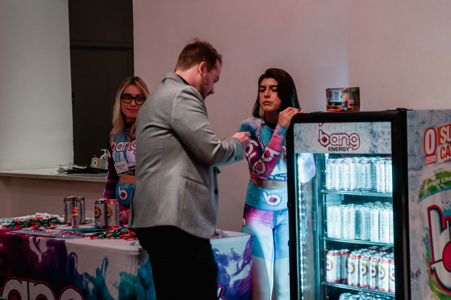 A man in a gray blazer talks to two women in colorful Bang outfits beside a glowing cooler of Bang cans.