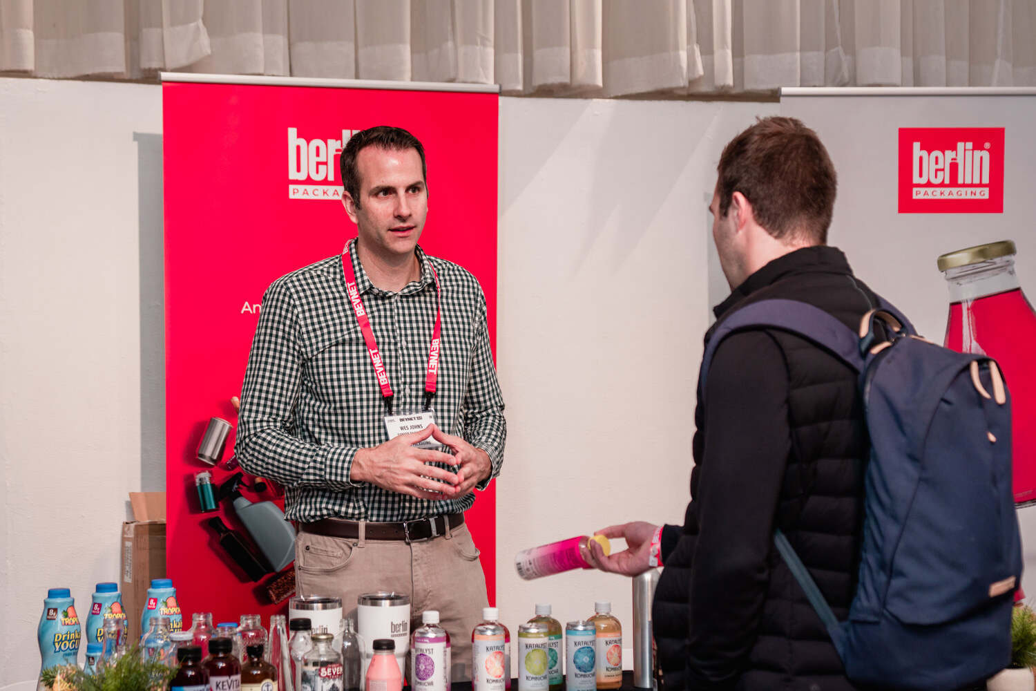 A man in a checked shirt gestures while an attendee with a backpack listens, with bottles on the table in front.