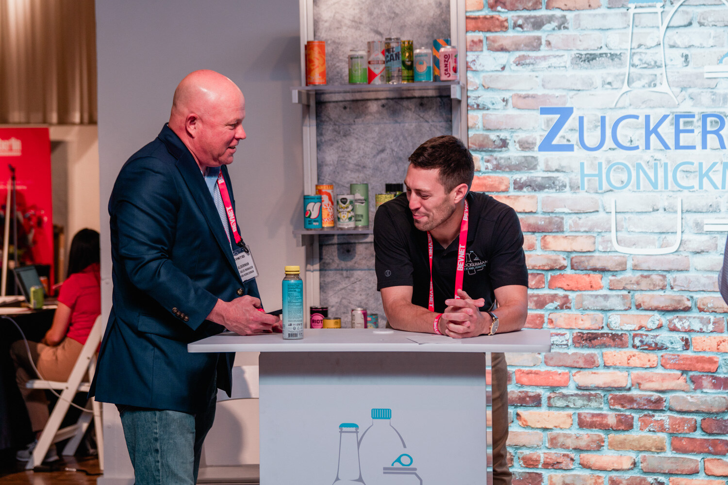 A bald man in a blue blazer and a man in a black polo lean on a white table in front of a brick wall display.