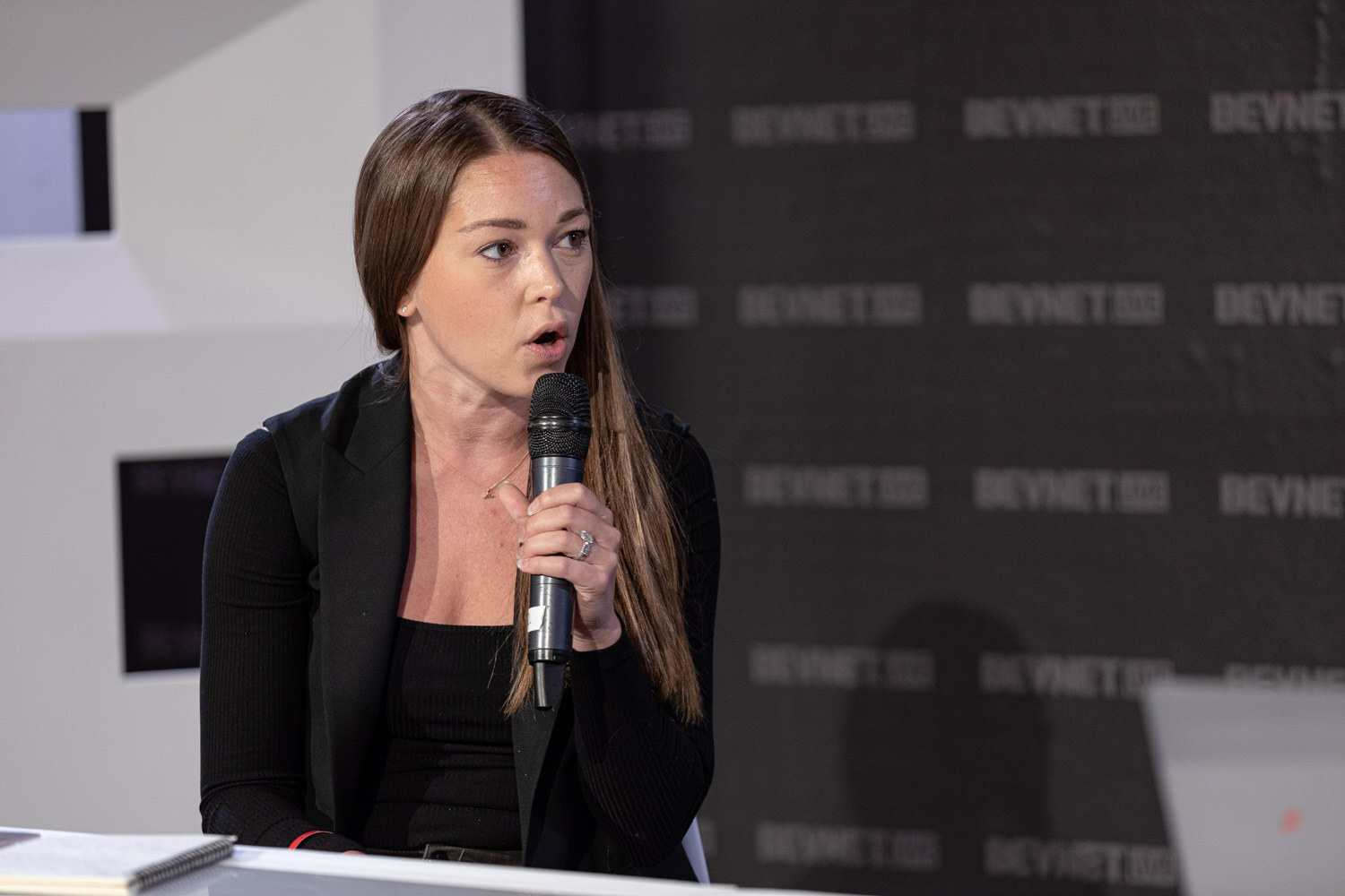 A woman with long brown hair in a black blazer holds a microphone and looks off to her left against the BevNET Live backdrop.
