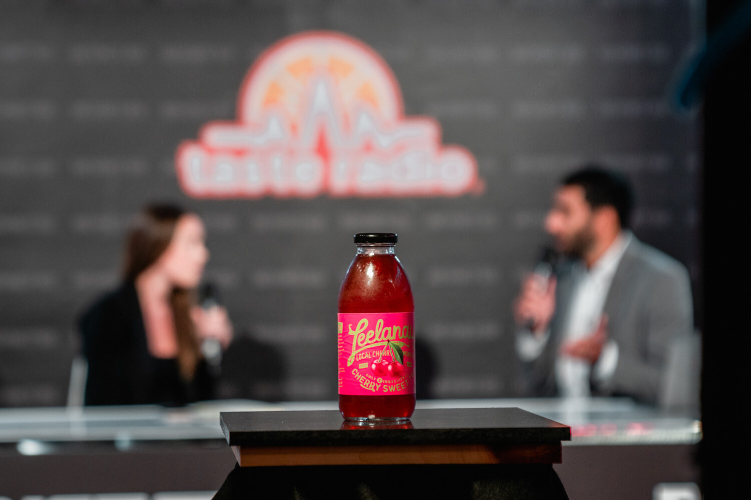 A bottle labeled Leelanau Cherry Sweet in focus in front of two blurred people at the Taste Radio livestream desk.