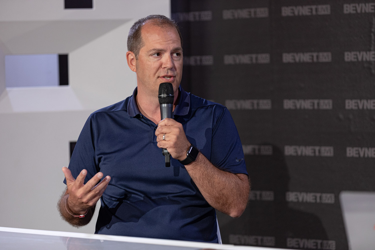 A man in a navy polo holds a microphone with one hand and gestures with the other in front of the dark backdrop.