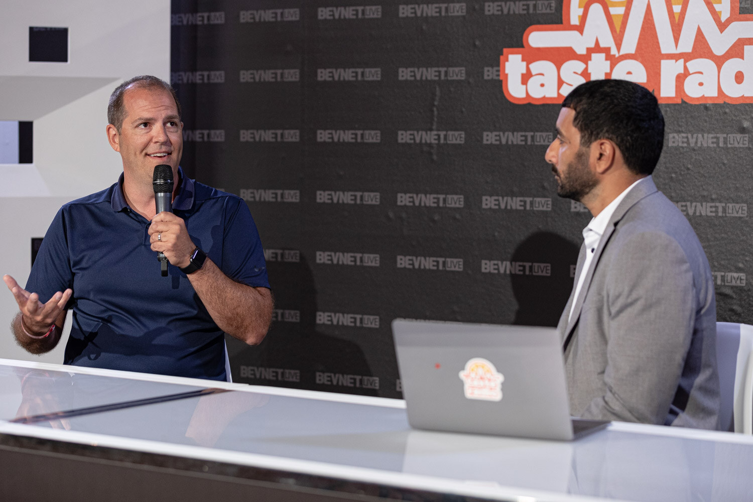 A man in a navy polo holding a microphone gestures to a man in a gray blazer who sits beside a laptop with a Taste Radio sticker.