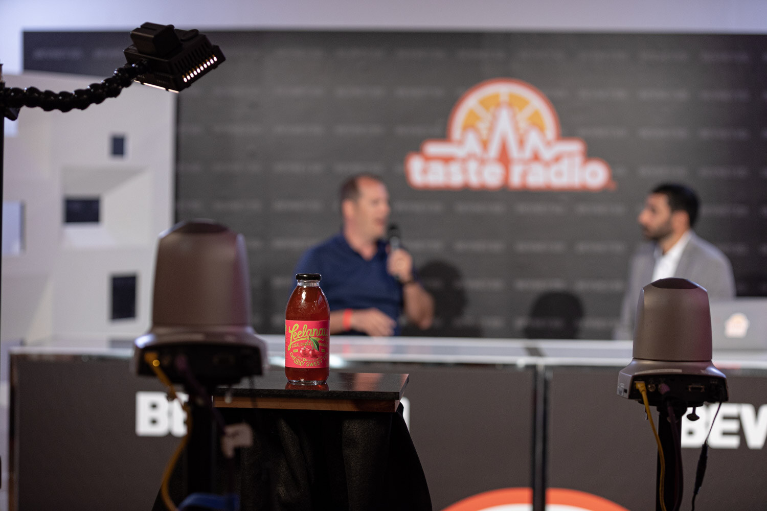 A bottle labeled Leelanau Cherry Sweet in the foreground with two broadcast cameras, and two people talking at a white desk under the Taste Radio sign.
