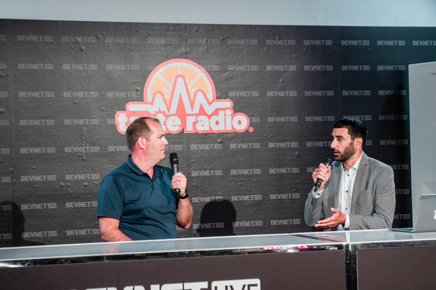 A man in a teal polo and a man in a gray blazer hold microphones and talk at a white desk in front of a Taste Radio logo wall.