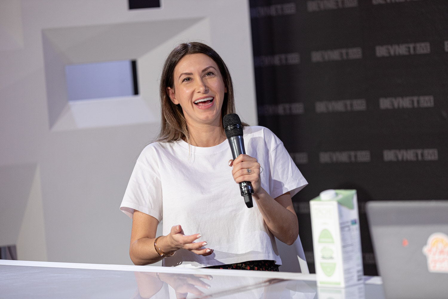 A woman with long brown hair and a white t-shirt holds a microphone and smiles at the desk, with a green carton beside her.