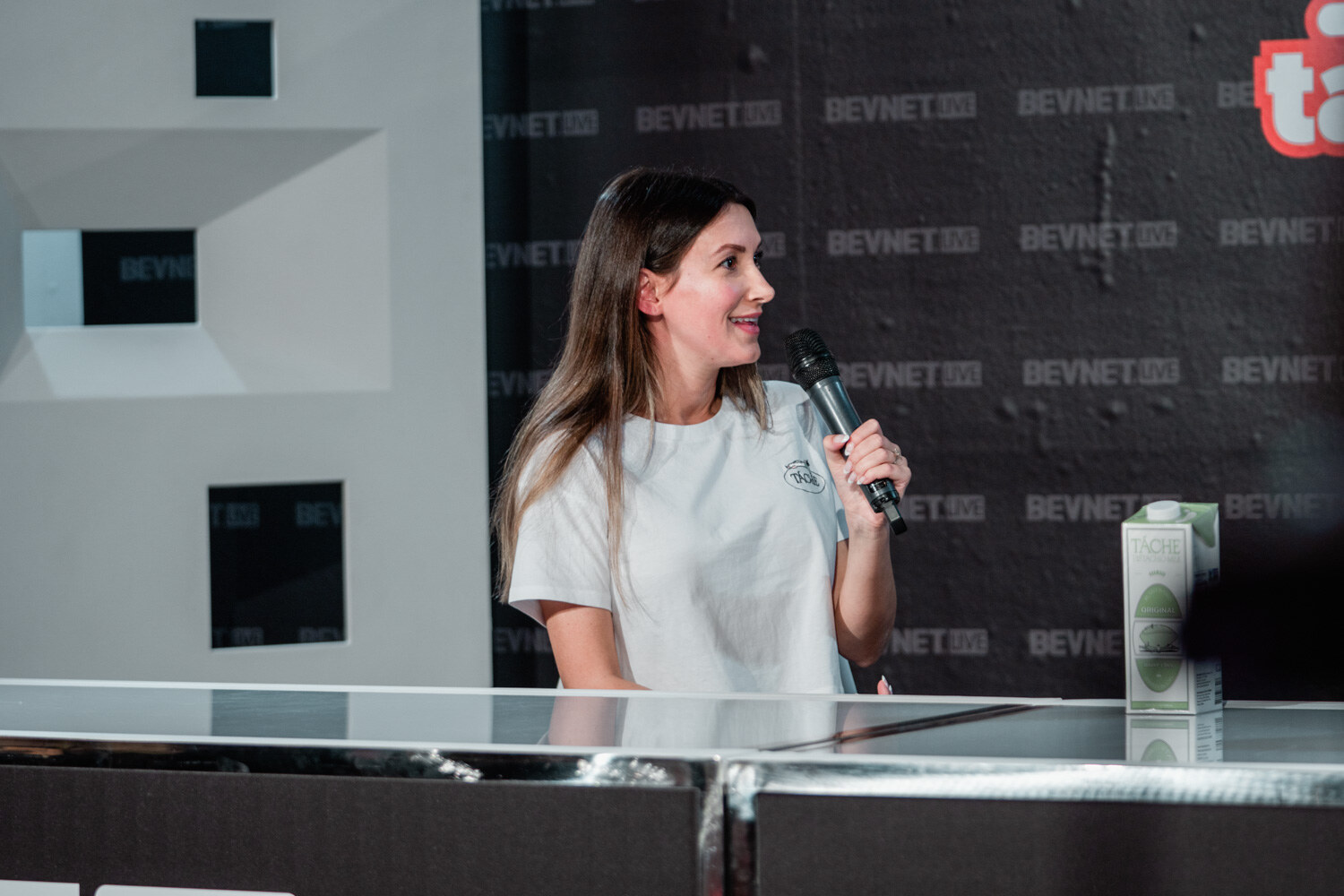 A woman with long dark hair and a white t-shirt speaks into a microphone in front of a dark BevNET Live backdrop with a carton of Tache beside her.