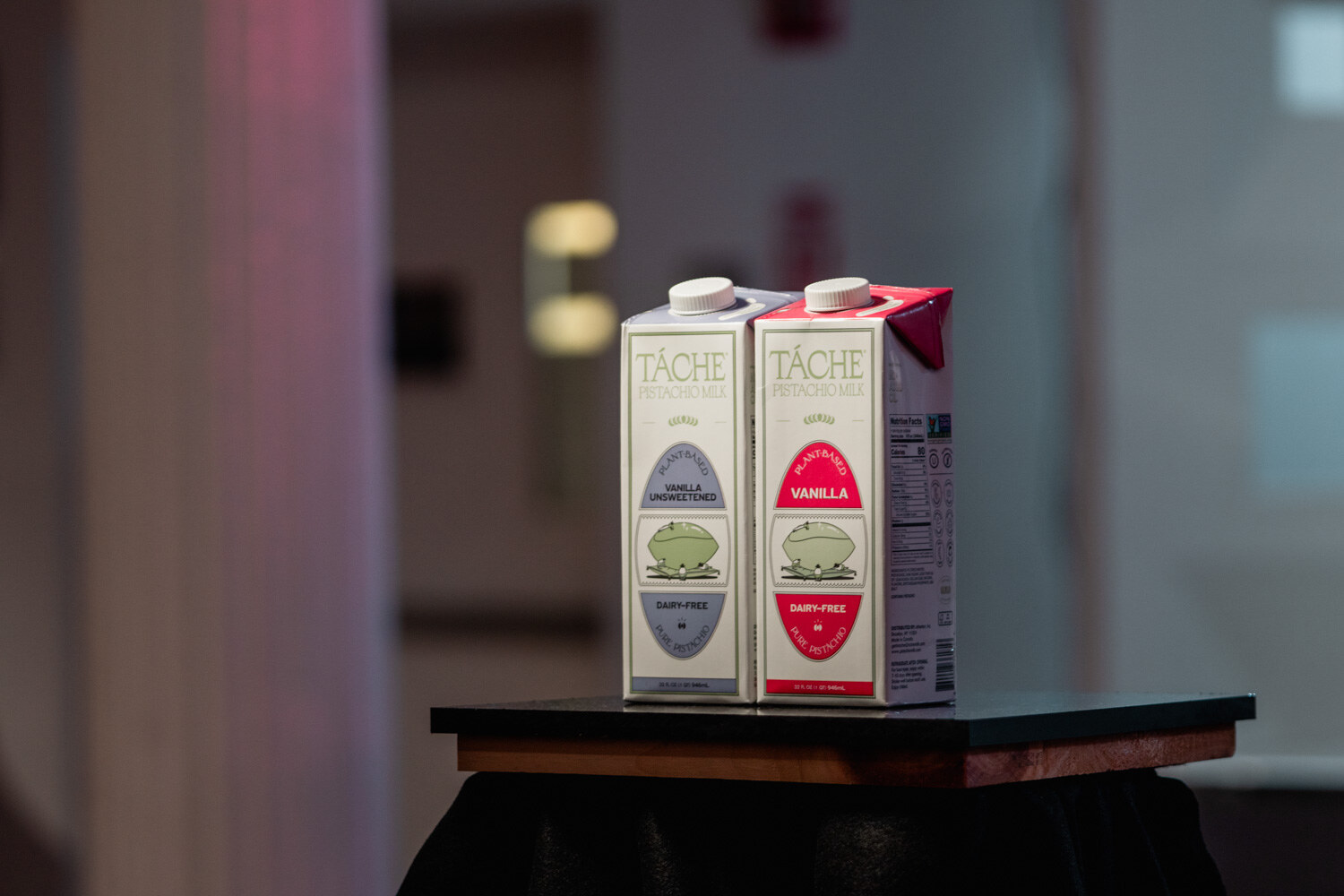 Two cartons of Tache pistachio milk, one with a blue label and one with a red label, on a small black table.