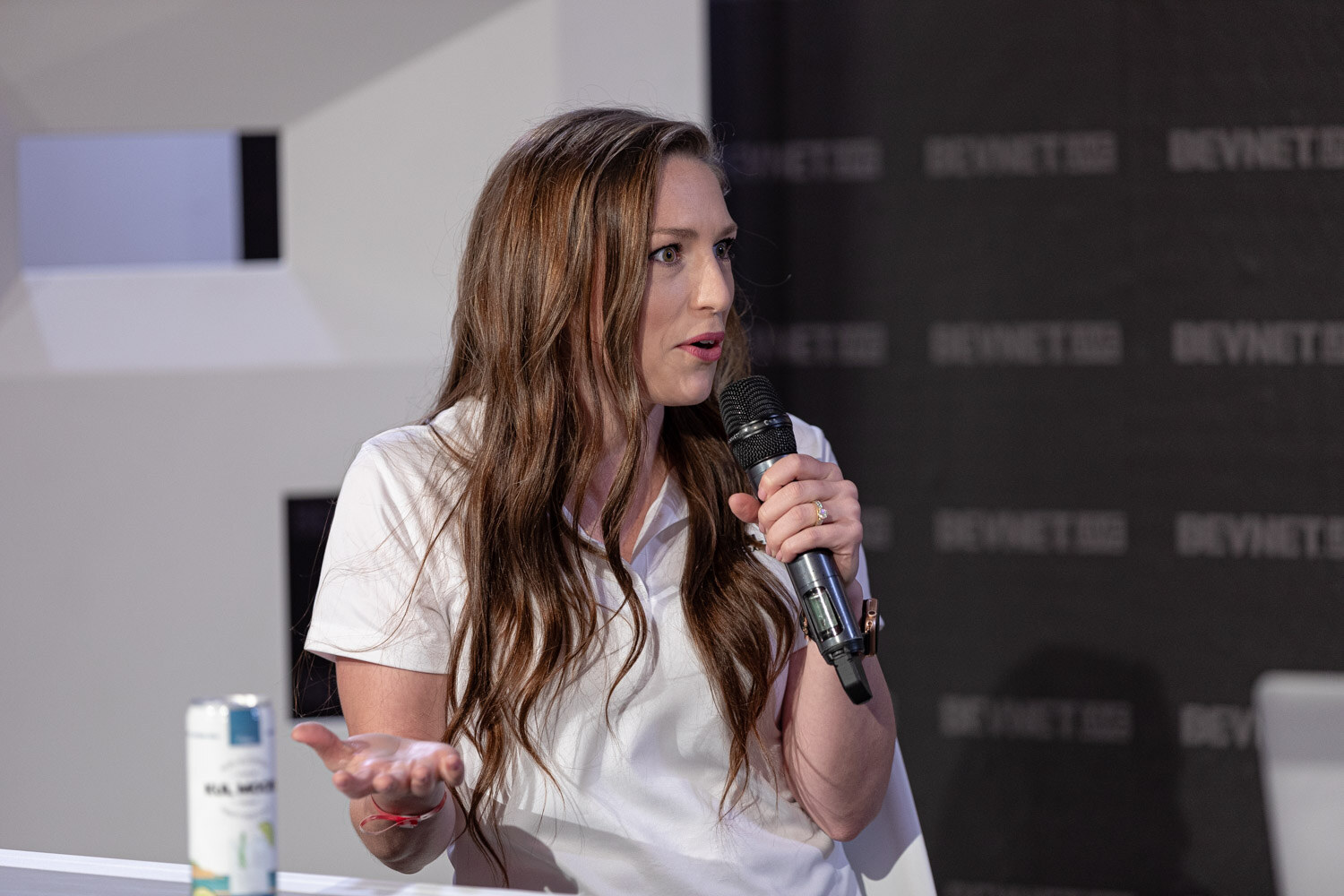 A woman with long wavy hair and a white shirt holds a microphone and gestures with her other hand, a can on the desk.