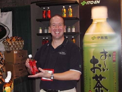 A smiling bald man in a dark polo holds a red-labeled bottle in front of shelves of bottled teas and a large Ito En green tea bottle graphic with Japanese text.