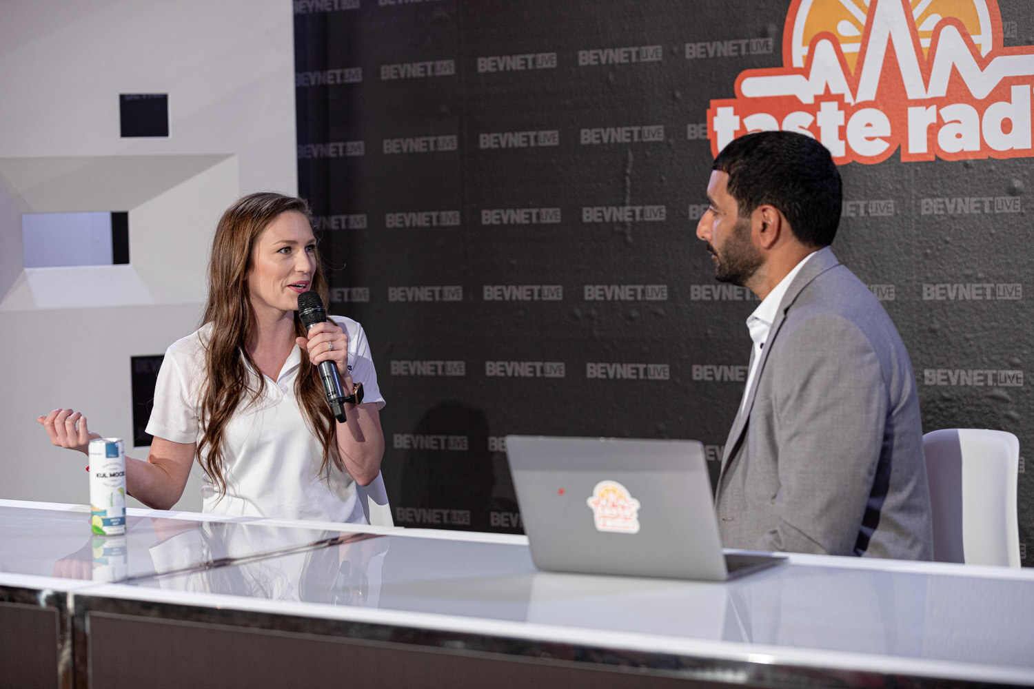 A woman in a white polo with a microphone talks to a man in a gray blazer at a white desk with a laptop and a can.