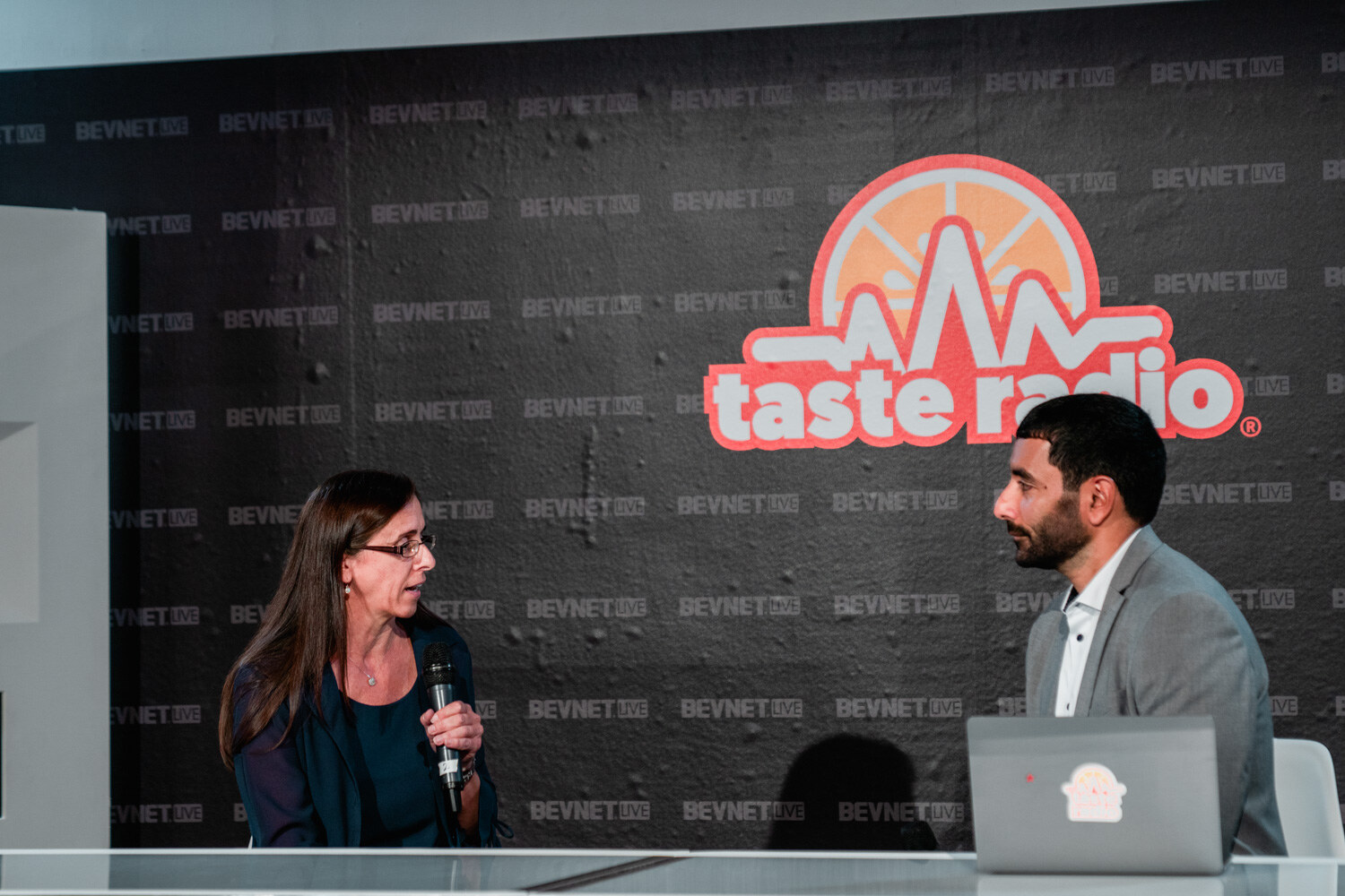 A woman holding a microphone and a man in a gray blazer sit at a desk under a large Taste Radio logo, a laptop in front of the man.