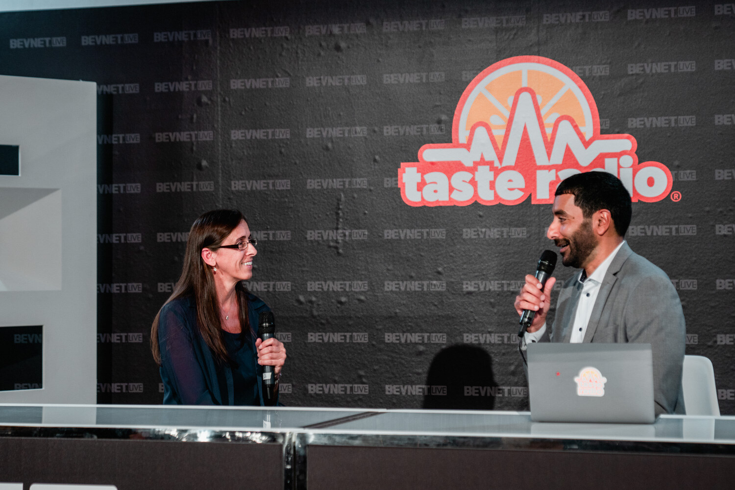 A man in a gray blazer holding a microphone talks with a woman with glasses and long brown hair at a white desk under the Taste Radio sign.
