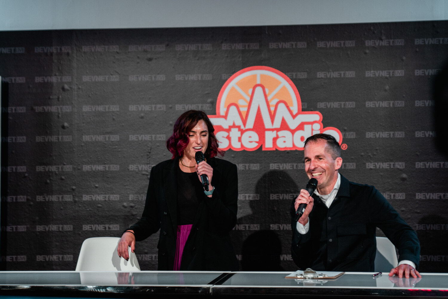 A woman with burgundy hair and a man in a dark jacket hold microphones and smile at a white desk beneath the Taste Radio logo.