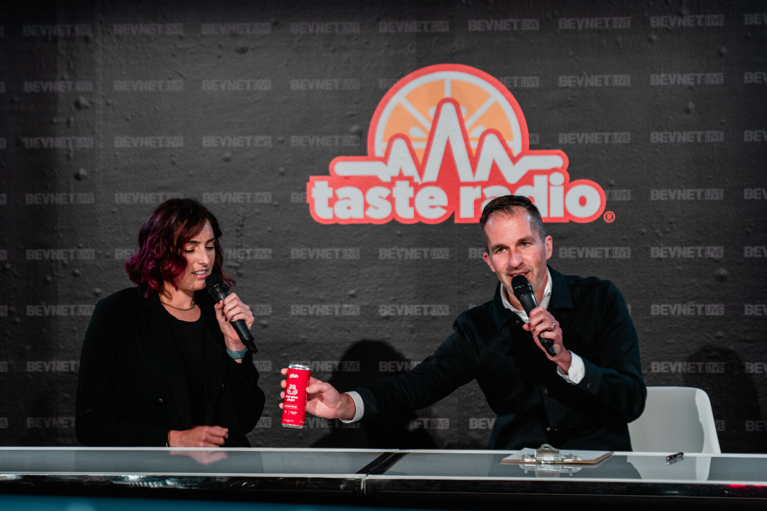 A man in a dark jacket holds out a red can toward a woman with burgundy hair at a desk in front of the Taste Radio logo.
