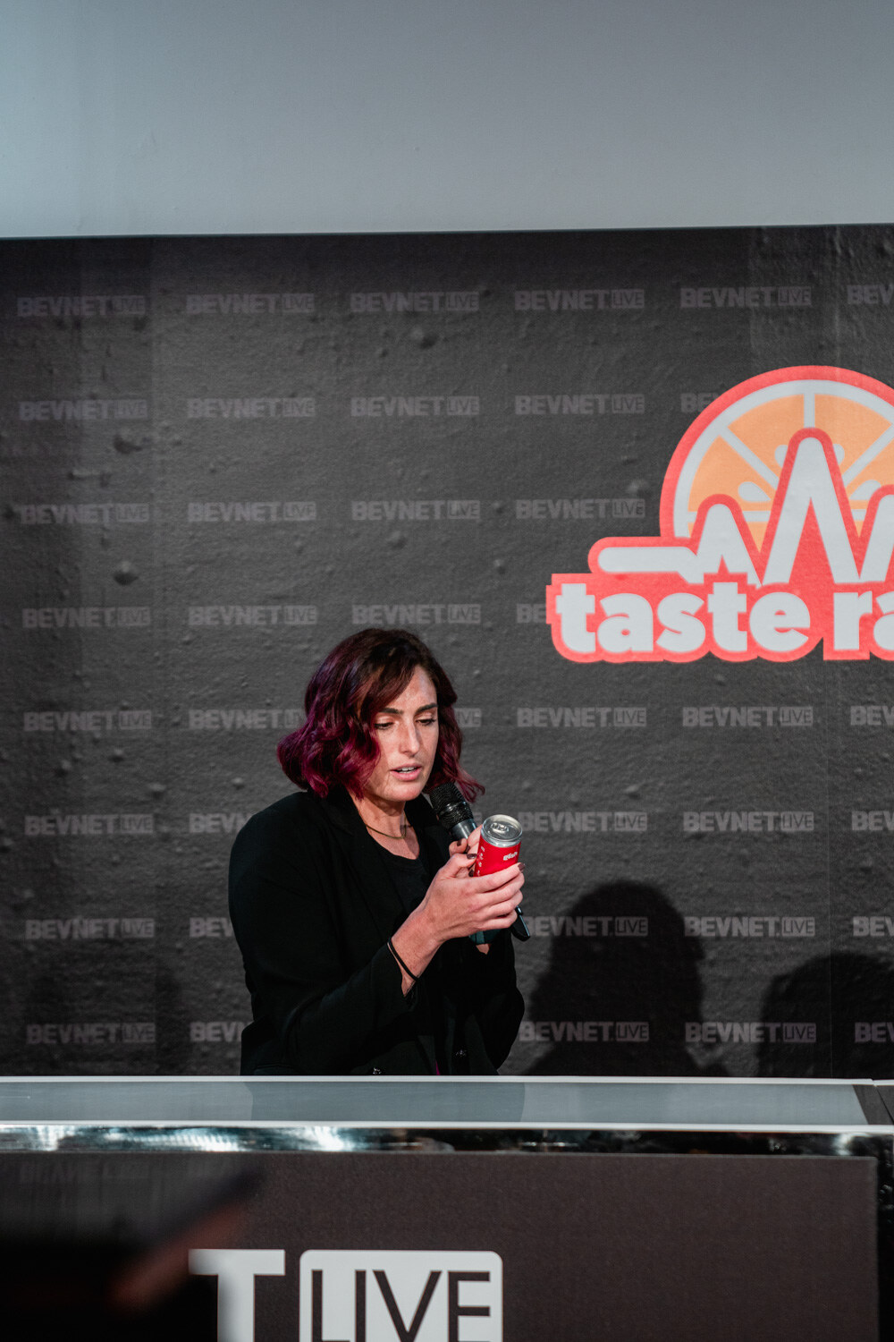 A woman in a black jacket with burgundy hair sits at the desk holding a red can in front of her face, the Taste Radio logo behind her.
