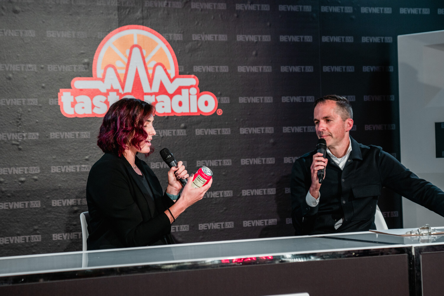 A woman with burgundy hair holds a red can toward the camera while a man in a dark jacket speaks into a microphone beside her.