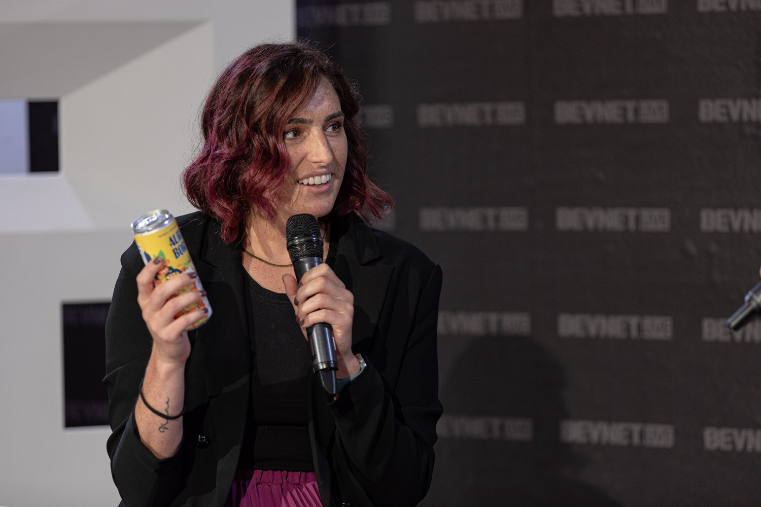 A woman with burgundy hair in a black blazer holds a yellow can in one hand and a microphone in the other.