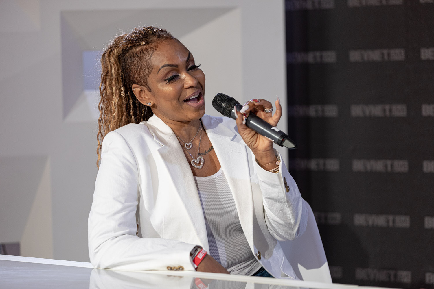 A woman with golden braided hair in a white blazer and heart necklace holds a microphone and smiles at the desk.