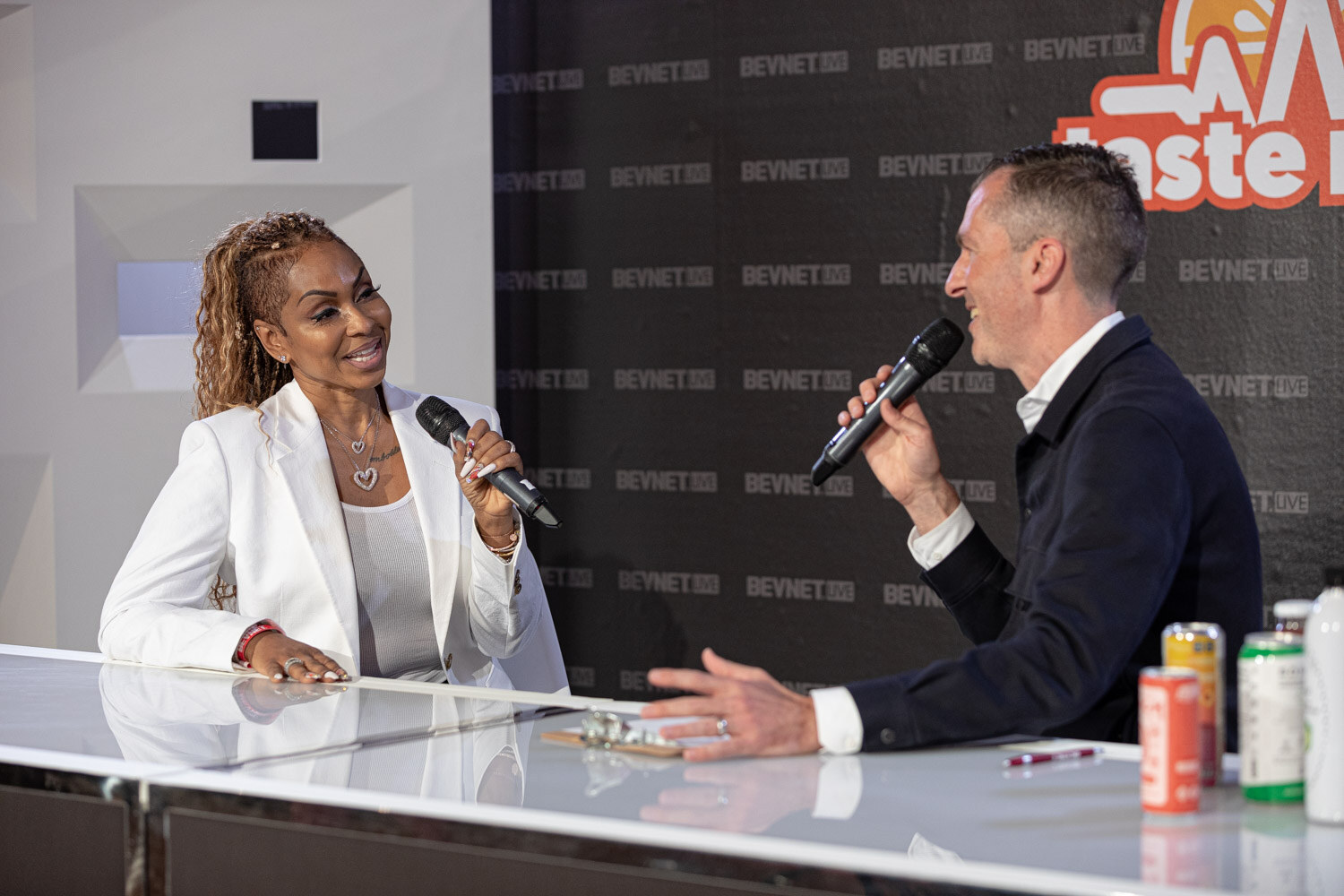 A woman in a white blazer and a man in a navy jacket hold microphones and talk across a white desk with several cans.