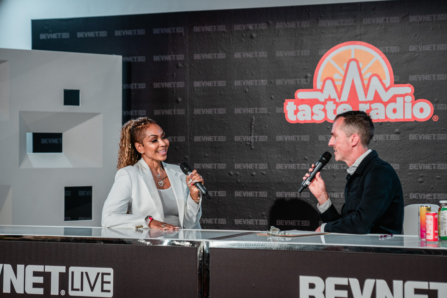 A woman in a white blazer holds a microphone and a man in a dark jacket speaks into one, with drinks at the right of the desk.