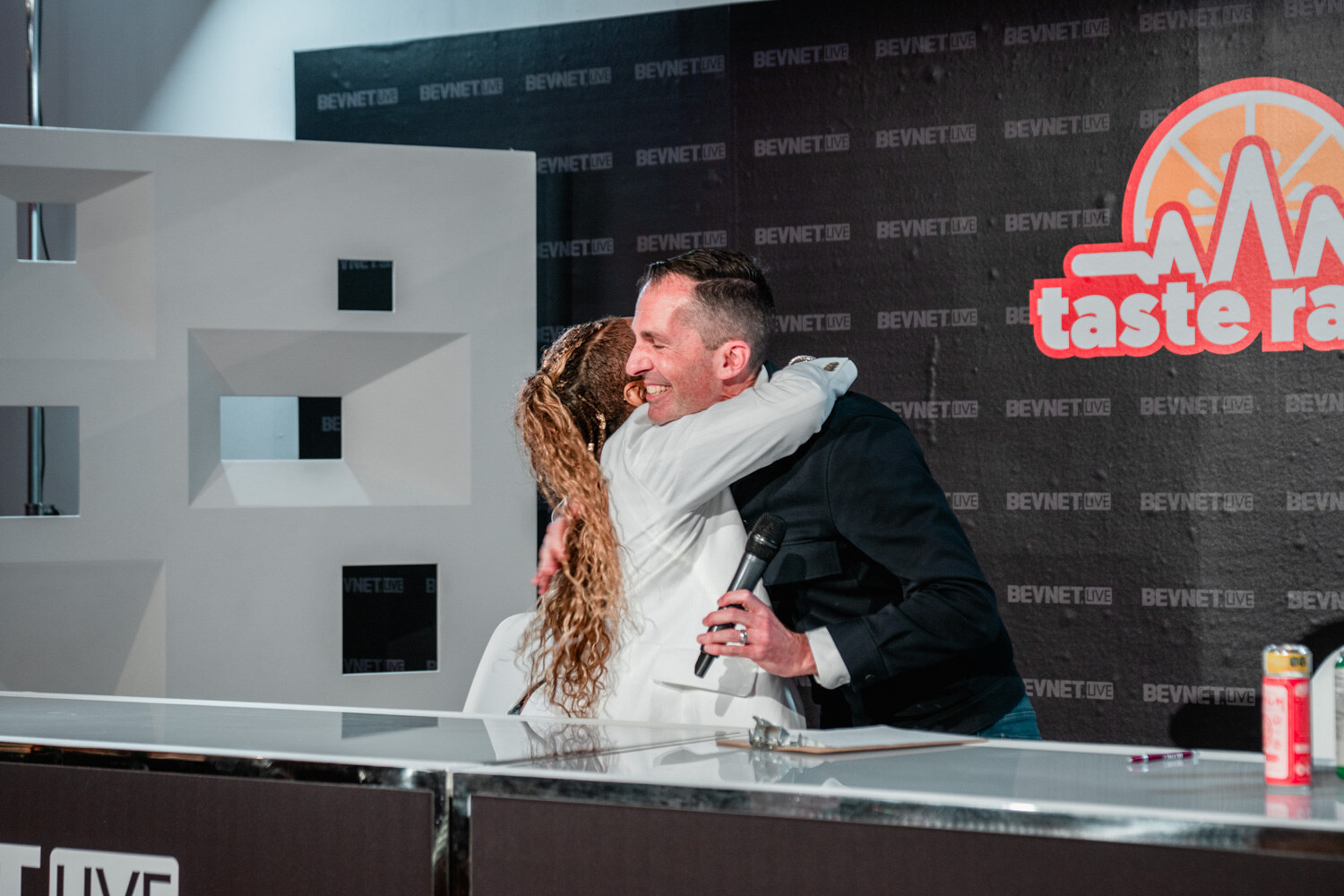A woman with long curly golden hair in a white blazer hugs a man in a dark jacket in front of the Taste Radio logo.