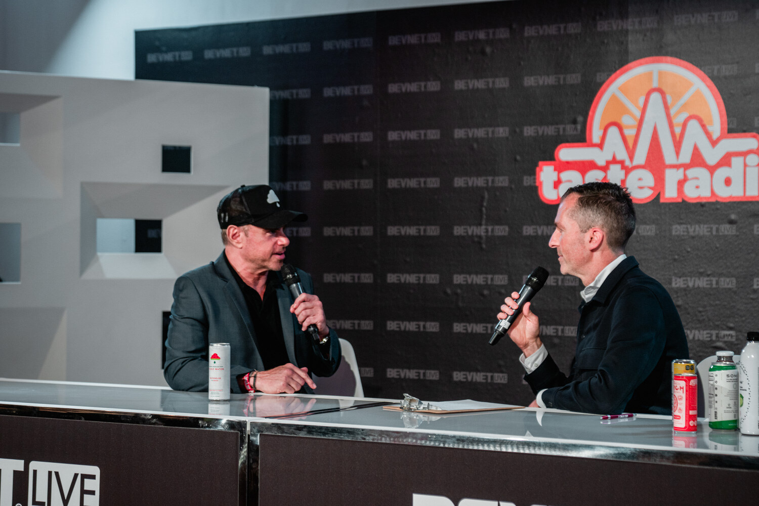 A man in a black cap and gray blazer holds a microphone and talks with a man in a dark jacket holding a microphone, cans on the desk.