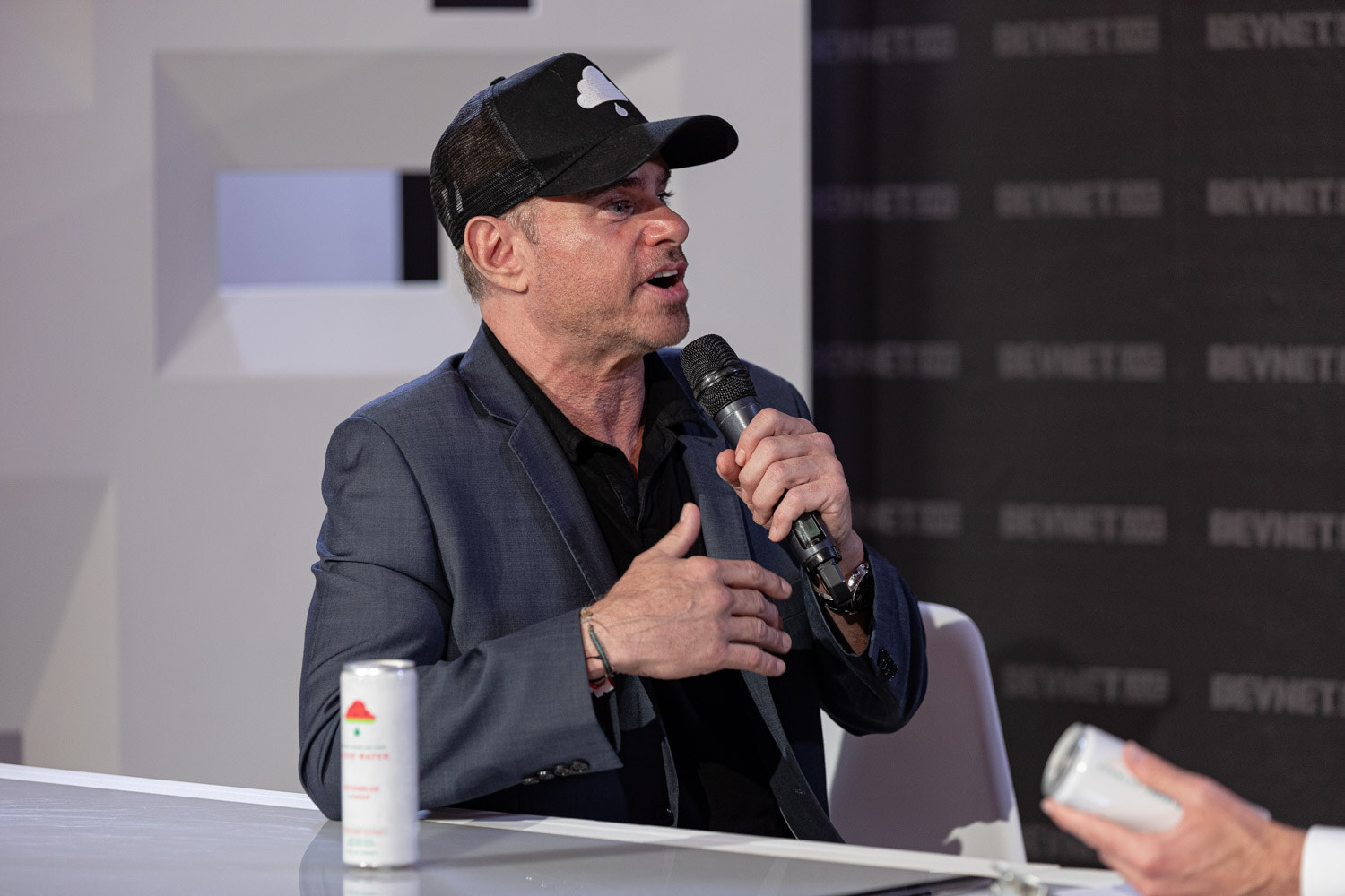 A man in a black cap and gray blazer holds a microphone and gestures at his chest, with a can on the desk beside him.