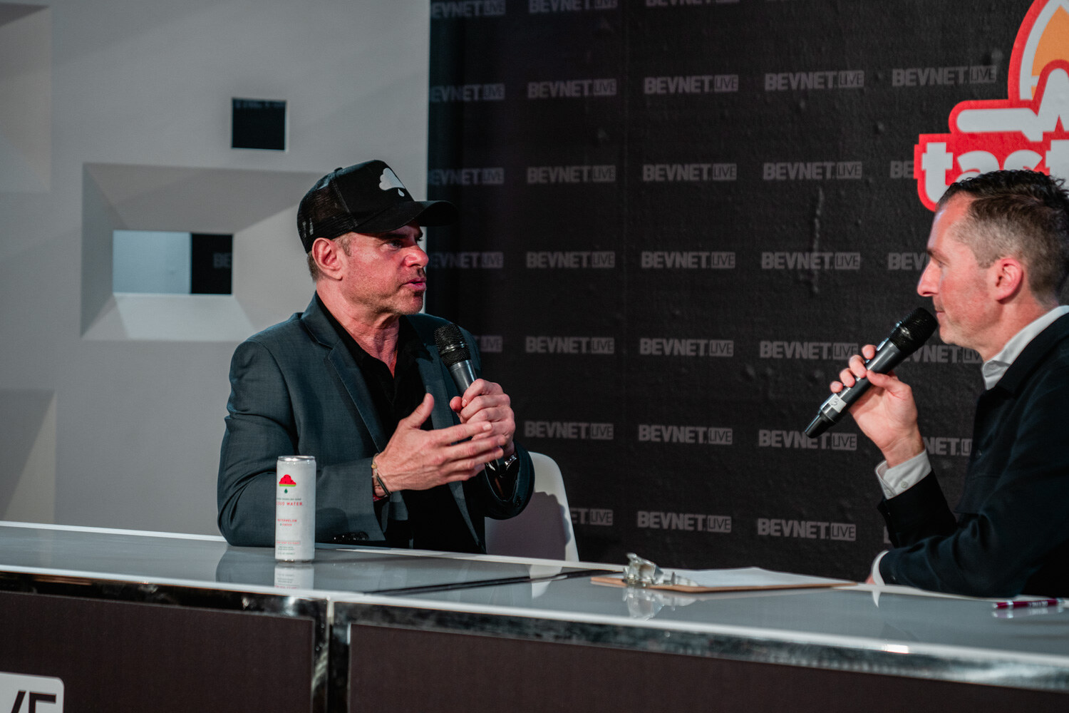 A man in a black cap and gray blazer gestures with one hand, a can in front of him, while a man in a dark jacket holds a microphone.