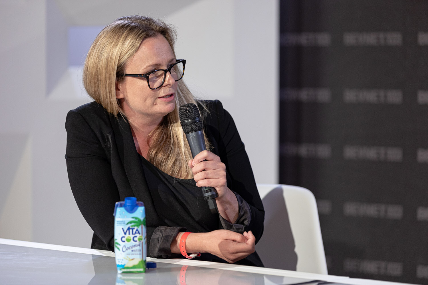 A woman with blonde hair and glasses in a black blazer holds a microphone with a carton of Vita Coco on the desk.