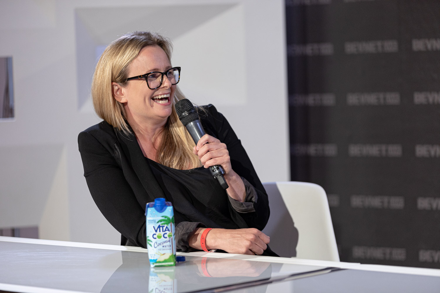 A woman with glasses and a black blazer laughs while holding a microphone, a Vita Coco carton on the desk.