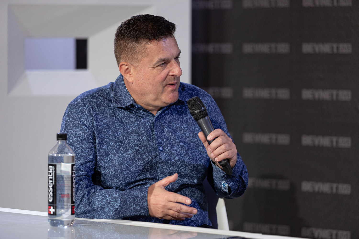 A man in a blue paisley shirt holds a microphone and gestures with one hand, with an Essentia water bottle on the desk.
