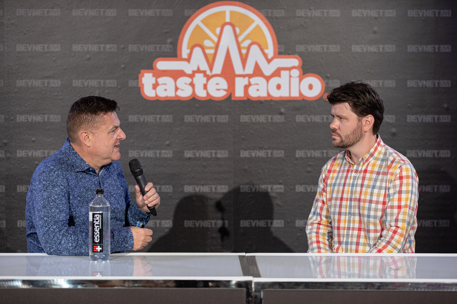 A man in a blue shirt with a microphone talks to a man in a multicolor plaid shirt at a desk with an Essentia water bottle.