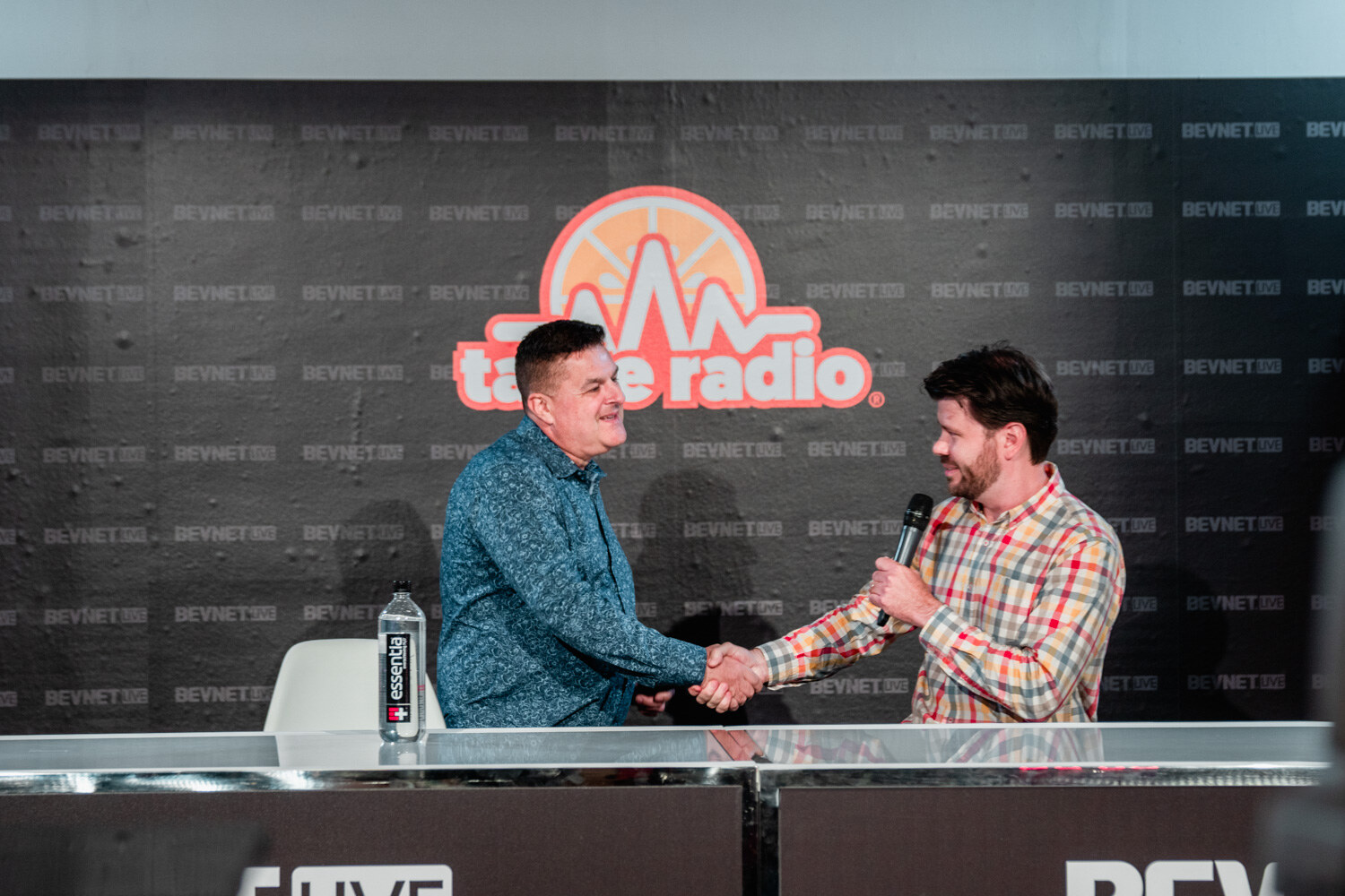 A man in a teal shirt and a man in a plaid shirt shake hands across a white desk in front of the Taste Radio logo.