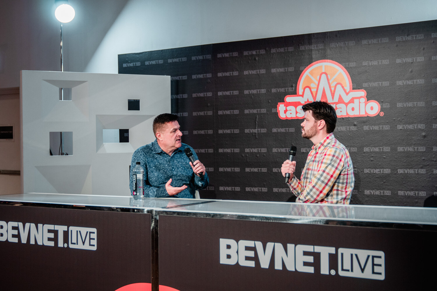 Wide view of two men talking at a white desk with a BevNET Live front panel, beneath the Taste Radio logo.