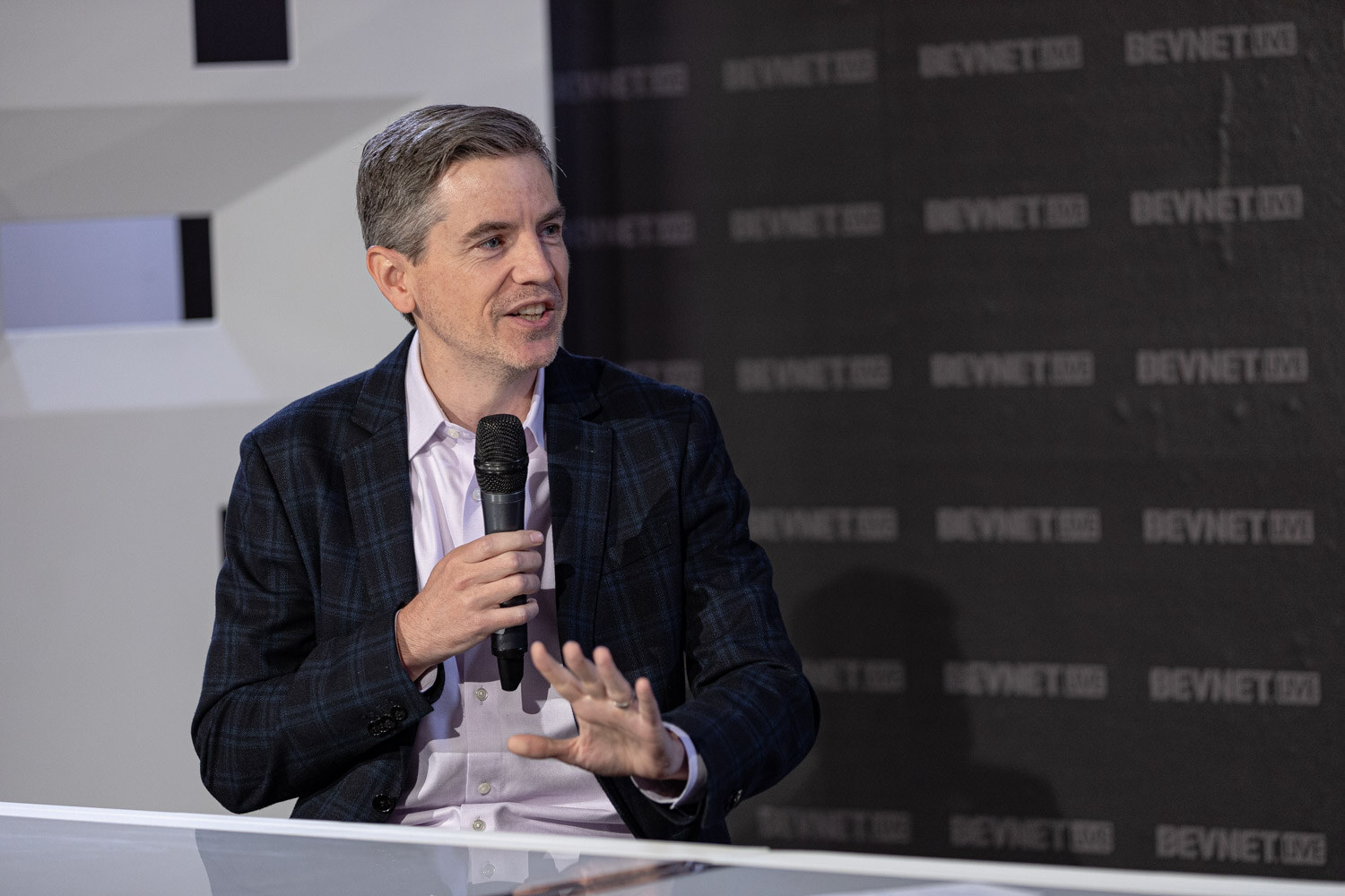A man with gray hair and a plaid blazer holds a microphone and gestures with his other hand.