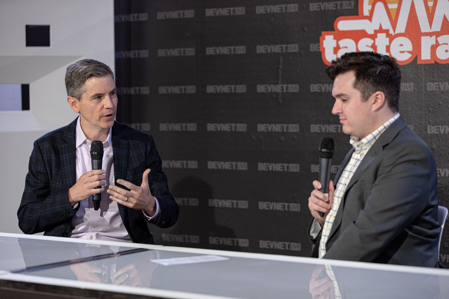 A man in a plaid blazer and a man in a gray blazer with a checked shirt hold microphones at a white desk with a dark backdrop.