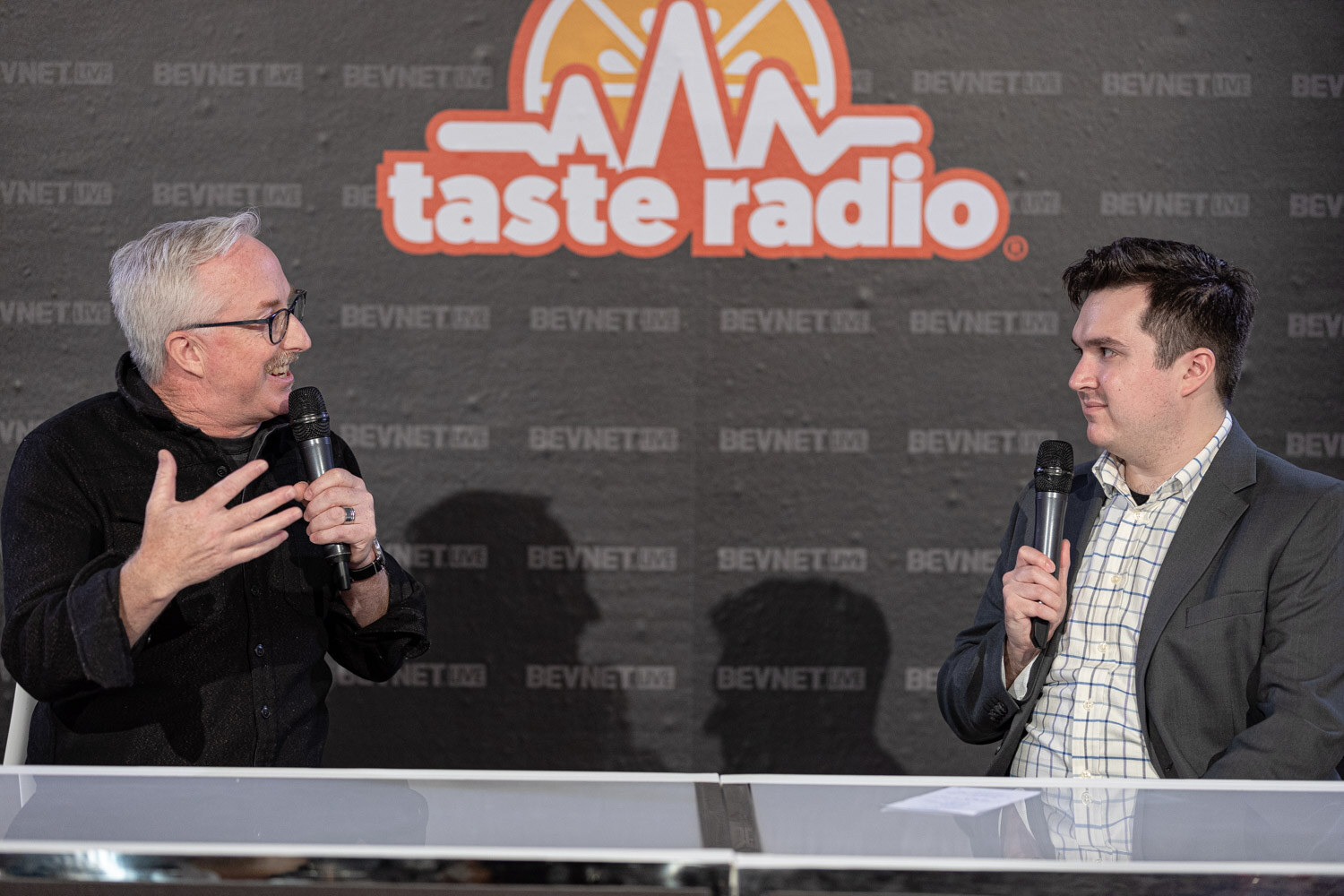 A man with gray hair and glasses in a black jacket smiles and gestures, facing a man in a gray blazer holding a microphone.