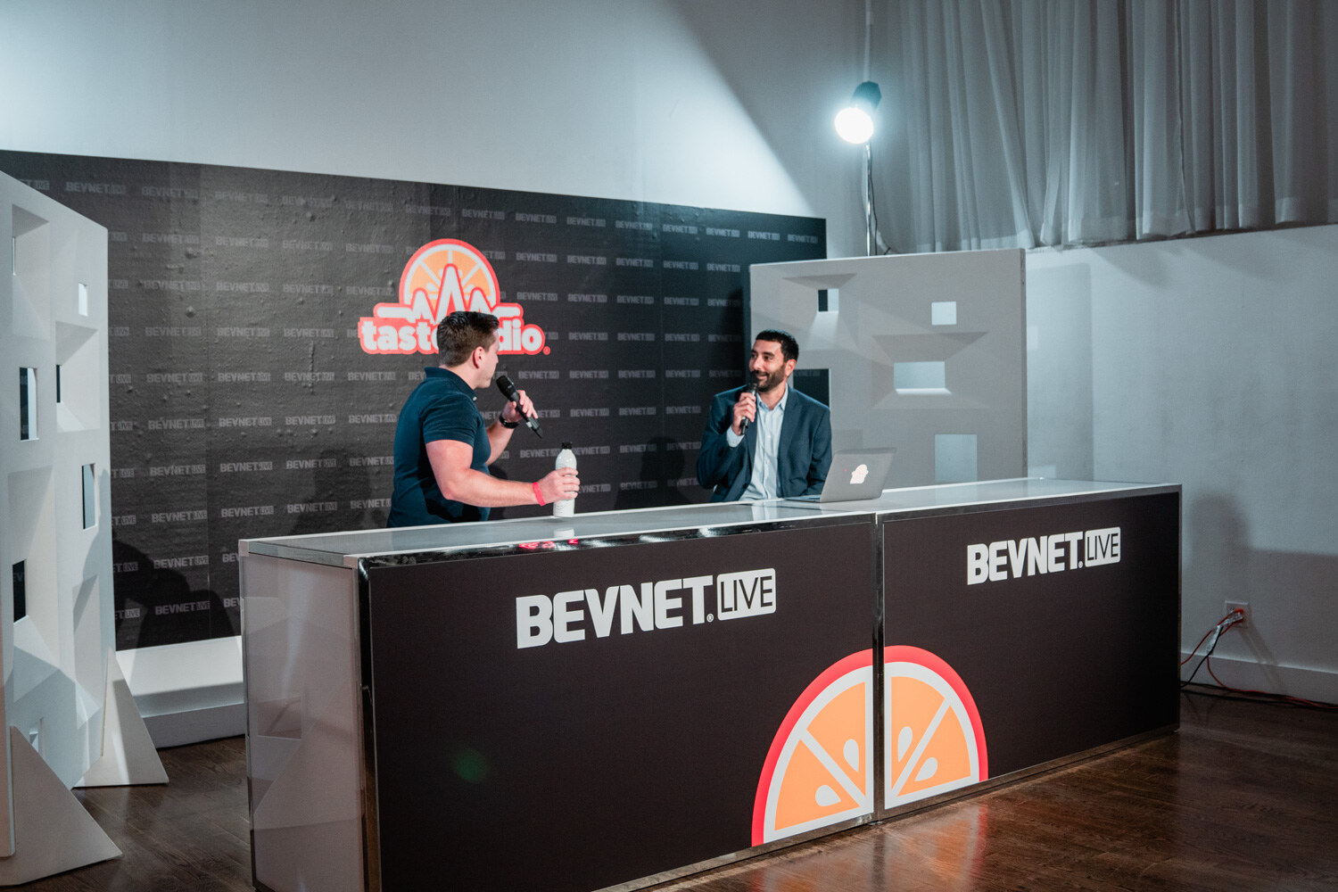 Wide view of the Taste Radio studio set with a desk branded BevNET Live and a Taste Radio logo wall behind two men talking.