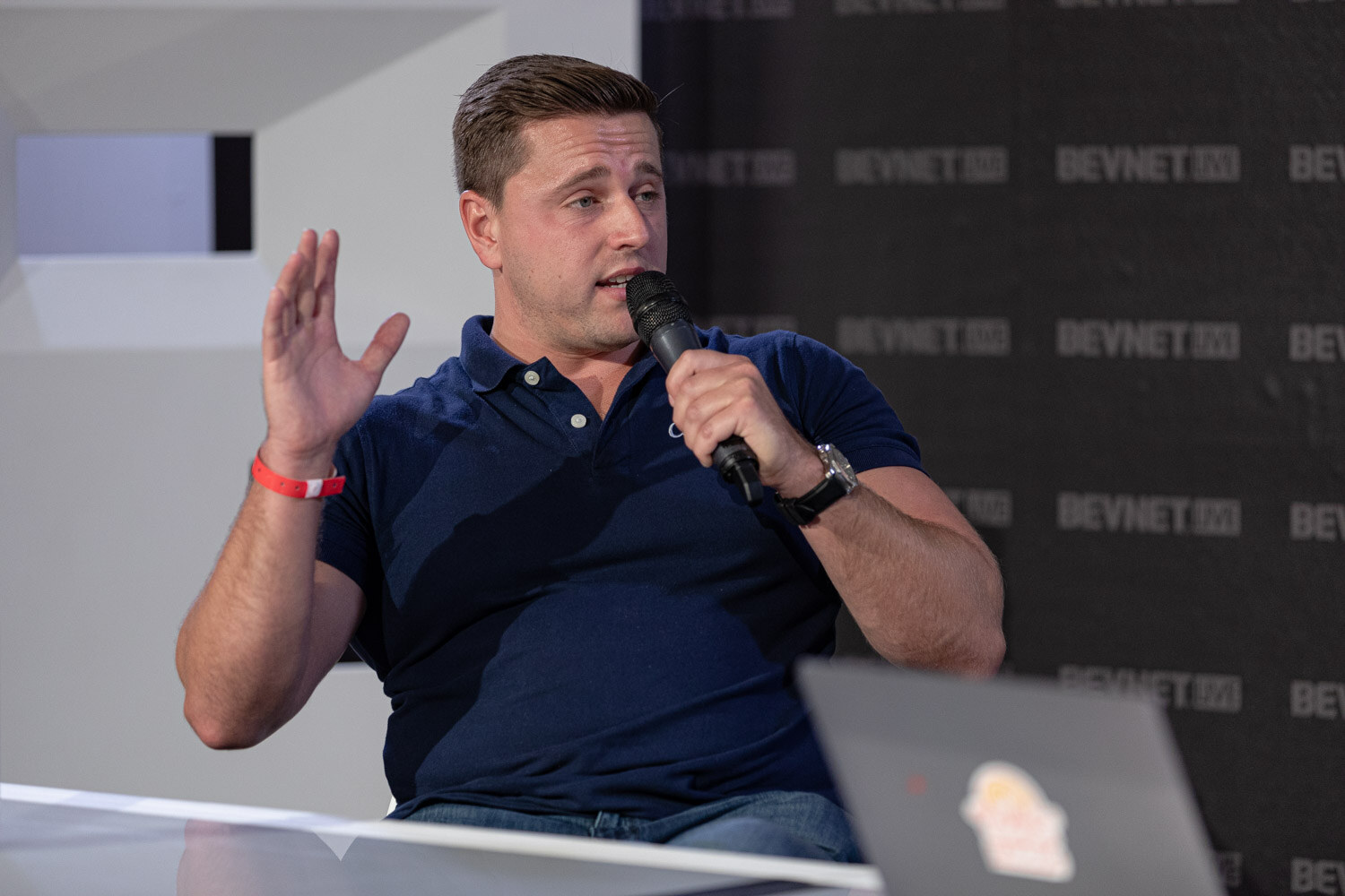 A man in a navy polo and a red wristband holds a microphone and raises one hand, a laptop at the lower right.