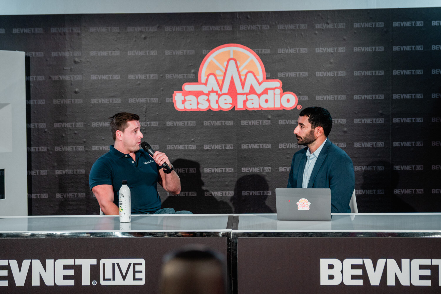 A muscular man in a teal polo with a microphone talks to a man in a gray blazer who sits at a laptop.